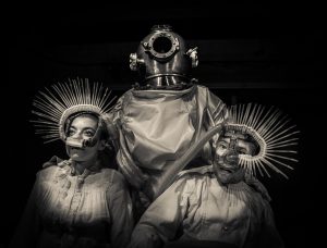  [Warm toned Black and white photo of 3 figures in surreal costumes. The central figure supports the two flanking figures. All are seated. The central figure wears a 19th century diving bell and a 20th century space suit. The flanking figures wear 19th century linen night shirts and strange breathing masks over their noses. Many spines protrude from each breathing mask, part medical device part undersea creature.]