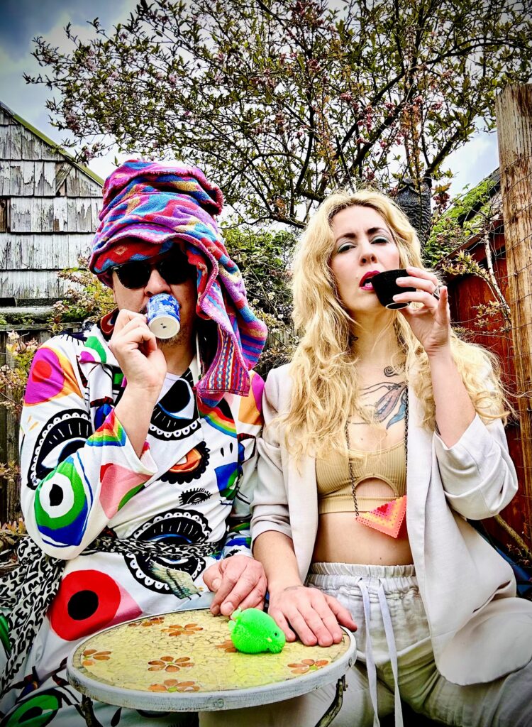 Two people in colorful costumes sip tea in a backyard