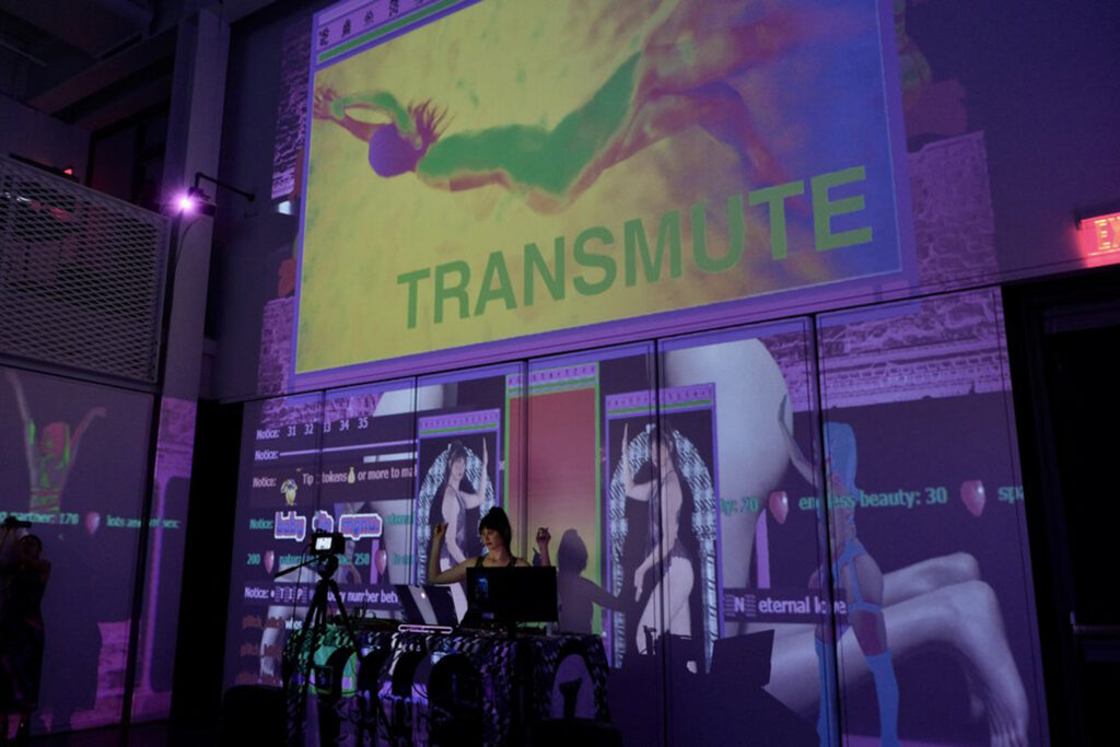 A DJ stands at a table in front of a large projection of psychedelic illustrations and the word TRANSMUTE
