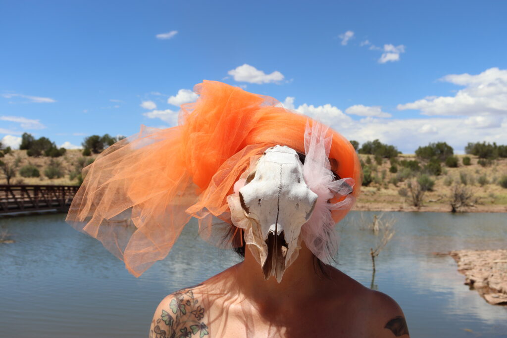 A naked figure stands in front of a river in an arid landscape, wearing a headdress of orange tulle atop a cow skull
