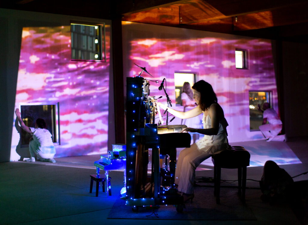 Photo of Jennifer Wright performing at a table with microphones and screens, with purple and pink light around her.